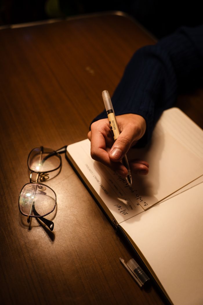 Close-up of a hand writing in a notebook with glasses on a wooden table.